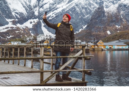 Traveler man taking self-portrait a photo with a smartphone. Tourist in yellow backpack standing on a background of a mountain and a lake a wooden pier.