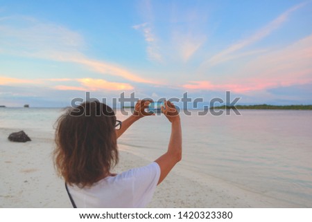 Woman taking photo with smartphone of romantic sky at sunset on sand beach, rear view, real people traveling around the world. Indonesia tropical destination, lifestyle sharing social media concept.