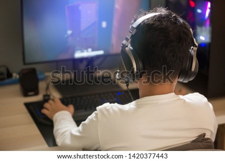 Portrait of asian boy gamer playing games on computer in the room at home, wearing headphones and using backlit colorful keyboard on wooden table.