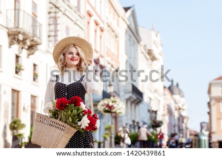 Outdoor portrait of young happy smiling lady wearing straw hat, white blouse, polka dot dress, holding straw wicker bag with flowers, posing in street of European city. Copy, empty space for text