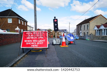 Digging the road up in a residential district to repair or replace water pipes with safety fencing, traffic cones and stop lights with a welsh language bilingual warning sign.
