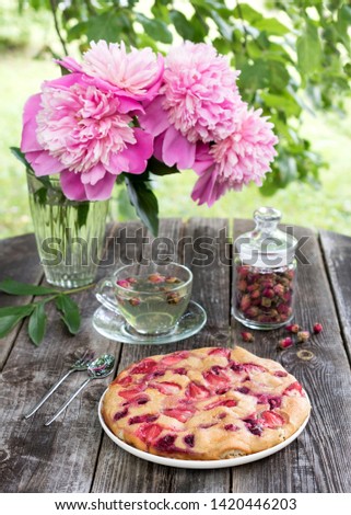 French dessert clafoutis with strawberries and cherries with tea in a cup of rosebuds. Peonies in a vase on a wooden background.