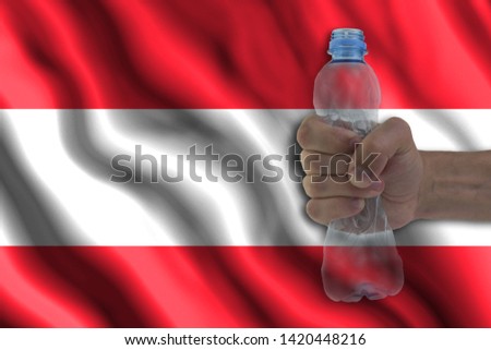 Concept of stopping plastic pollution. Ban disposable products. A hand squeezes a plastic bottle to protest the use of disposable plastic containers against the background of the flag of Austria