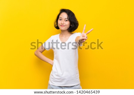 Asian young woman over isolated yellow wall smiling and showing victory sign
