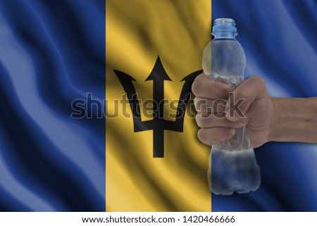 Concept of stopping plastic pollution. Ban disposable products. A hand squeezes a plastic bottle to protest the use of disposable plastic containers against the background of the flag of Barbados