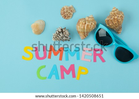 Text SUMMER CAMP of multicolored paper letters and sunglasses and seashells on a bright blue background. top view. flat lay