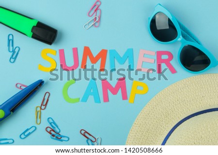 Text SUMMER CAMP of multicolored paper letters and sunglasses, hat and paperclip against a bright blue background. top view. flat lay