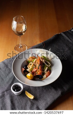 Seafood made dish with king prawns, mussels, squid, greens and citrus served with white wine and black olives on a white plate and wooden table