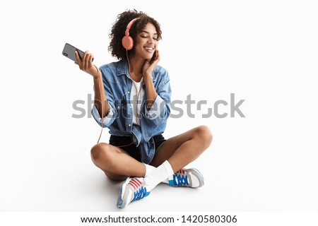 Full length portrait of a young cheerful african woman wearing casual denim clothes isolated over white background, listening to music headphones, holding mobile phone, dancing