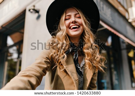 Cute blonde in a hat and a fashionable scarf around her neck makes a selfie. Portrait of a girl with burning cheerful eyes and an amazing smile.