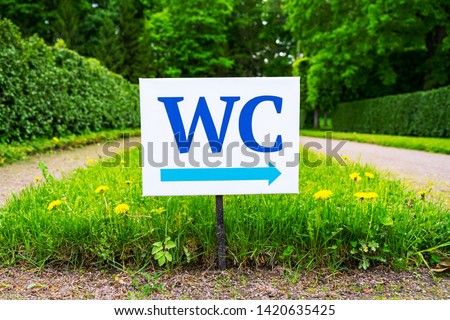 toilet sign against the background of green trees in the park.  White WC sign on white metal plate with blue pointing arrow indicating the direction