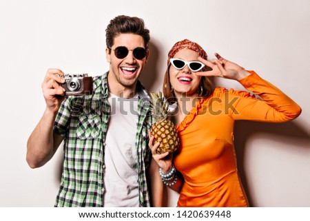 Portrait of brunet man taking picture on retro camera and woman in orange dress and headband. Girl in glasses holds pineapple and shows peace sign