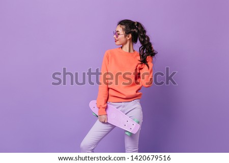 Confident curly korean girl holding longboard. Studio shot of charming asian model with skateboard isolated on purple background.