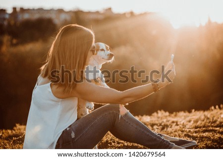 
Pretty young woman taking pictures with her sweet little dog with her smartphone on a sunny summer afternoon. Golden light. Lifestyle