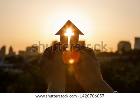 Paper house with cross in hand at sunset.