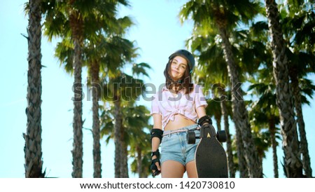A beautiful young brunette girl in a helmet and skate protection is standing in the park on the background of high palm trees, holding a skate in her hands, looking at the camera and smiling