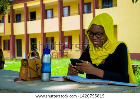 young beautiful black woman sitting down pressing her phone smiling with her book, water bottle and bag on the table