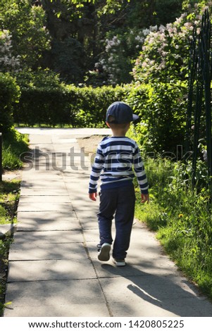European boy walks  without his parents along a narrow path, lined with large stone tiles, in a flowered square.  goes to the intersection of asphalt roads in the park