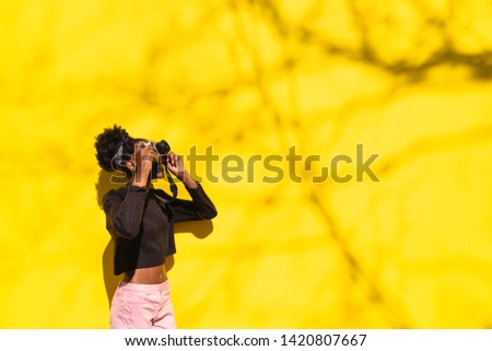 African smiling young woman in black knitted sweater, pink pants and bandana taking pictures on retro photo camera on the background of yellow wall on sunny day. Hobby, leisure and technology concept