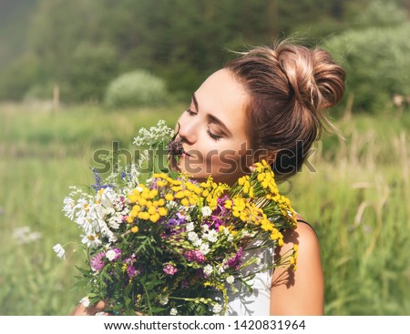 Summer lifestyle portrait  of beautiful young woman smiling and holding bouquet of wild flowers. Standing in the field of grass. Enjoying sun. Happiness and love concept. Сlosed eyes 