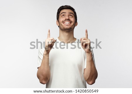 Closeup of European guy dressed in white T-shirt pointing upwards with positive face, isolated
