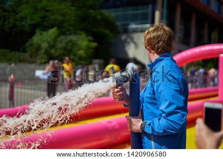 Man in the waterproof blue jacket pouring water from a hose onto a water slide during Bournemouth event. Dorset, England. Water sport and party background.
