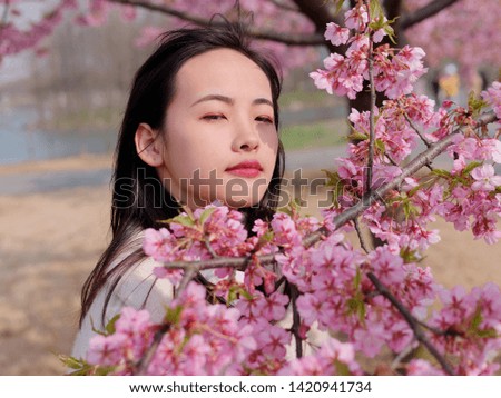 Head shot portrait of beautiful young brunette smiling woman behind pink cherry blossoms in sunny day. Outdoor portrait of Chinese stylish lady. Emotions, meditation, beauty and lifestyle concept.