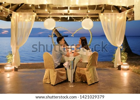 Young couple enjoying a romantic dinner by candlelight, outdoor.  Romantic meal on the beach with lanterns, chairs and decorations, sky, sea and beach in the background. Loving couple eating dinner. Royalty-Free Stock Photo #1421029379