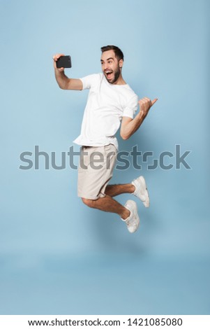 Full length photo of optimistic caucasian man in casual white t-shirt rejoicing while jumping and taking selfie on smartphone isolated over blue background
