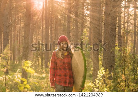 A happy surfer girl walking across a pine tree forest with a surfboard. Toned surfing lifestyle picture with cross light. Going for sunset surfing.