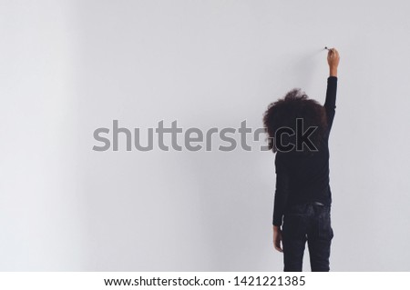 A dark-skinned girl is standing draws on a white wall.