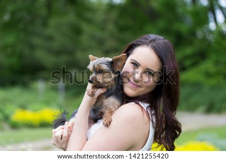 Horizontal medium shot of cute smiling brunette young woman in white top holding small Yorkshire Terrier dog seen in profile