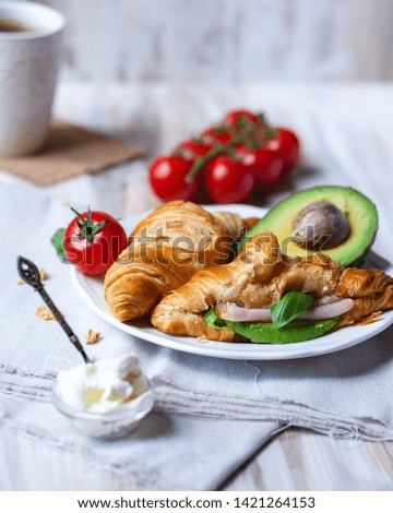 Delicious breakfast french style at home: light background, white plate with fresh croissants, small bowls of granola and cream cheese, cup of tea, avocado, tomatoes, basil. Served on beige napkins