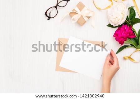 Top view woman hand holding wedding invitation card or love letter. Minimal flat lay composition with peony flowers, gift box and feminine accessories. Writing letter concept. 