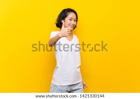 Asian young woman over isolated yellow wall with thumbs up because something good has happened