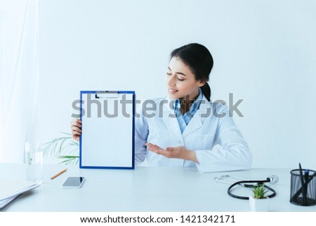 smiling latin doctor holding clipboard with blank paper while sitting at desk
