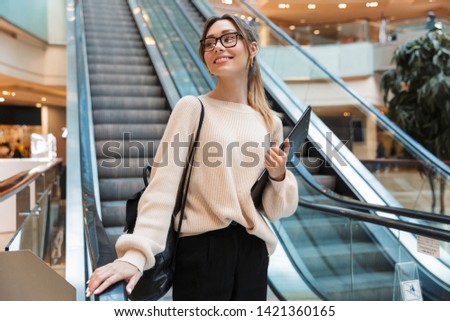 Photo of cute young woman wearing eyeglasses smiling and holding clipboard while going down escalator in building indoors