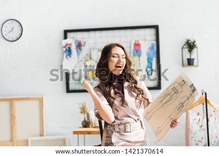 cheerful young designer gesturing while celebrating and holding fashion sketches 