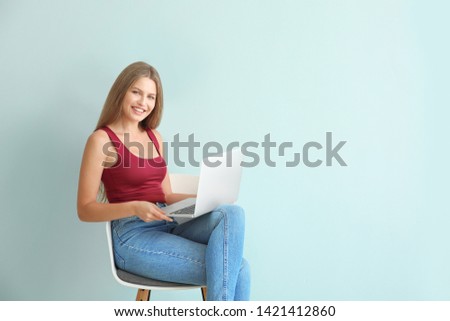 Beautiful young woman with laptop sitting on chair against color background