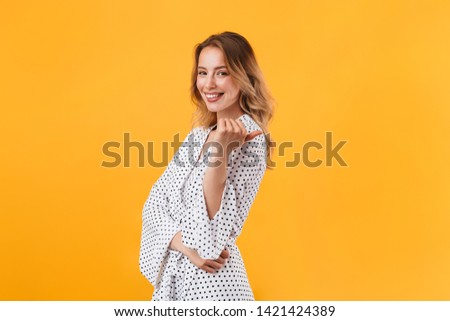 Beautiful young blonde woman wearing summer dress standing isolated over yellow background, pointing finger at copy space