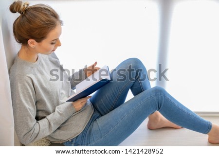 Beautiful young woman reading book near window