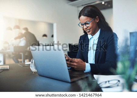 Smiling young African American businesswoman reading text messages and working on a laptop while sitting at her desk in a bright modern office