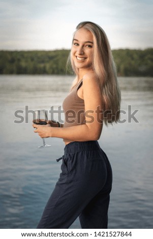 Pretty young woman in summer time with her red glass of wine in front of a calm lake at sunset