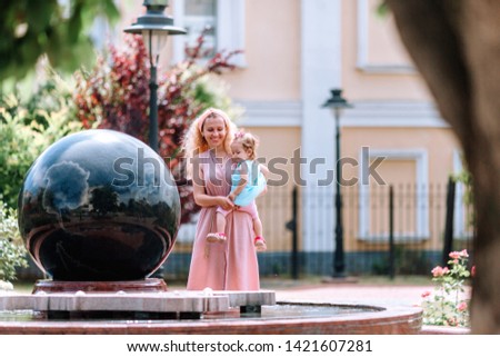 A happy blond woman dressed in a pink dress holds her little daughter in her arms, near the fountain in the form of a globe in a city park.