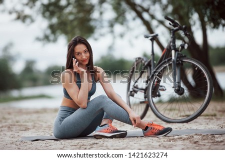 Making a call. Female cyclist with good body shape sitting near her bike on beach at daytime.