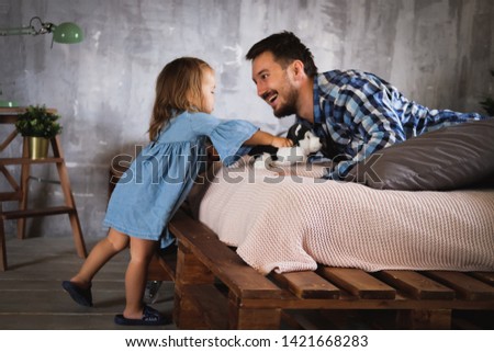 Dad and his daughter play on the bed in the bedroom plush dog. Daddy's baby. Dad with a beard in a plaid shirt, a girl in a denim dress. Happy family. Father's day