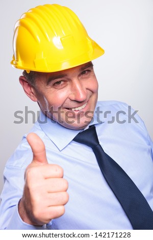 high angle picture of a senior bussines man wearing a helmet and showing thumb up and a smile at the camera.on gray background