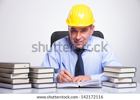 senior business man with helmet sitting at his desk, among piles of books and writing while smiling to the camera. on gray background