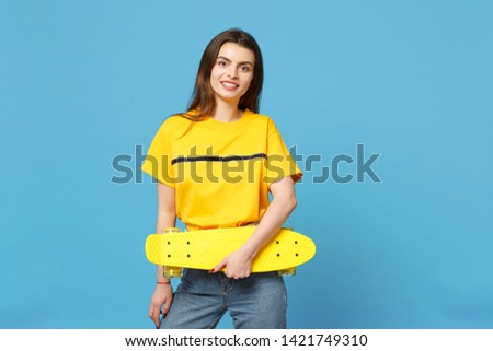 Portrait of cheerful young woman in vivid casual clothes standing, looking camera, holding yellow skateboard isolated on blue wall background in studio. People lifestyle concept. Mock up copy space