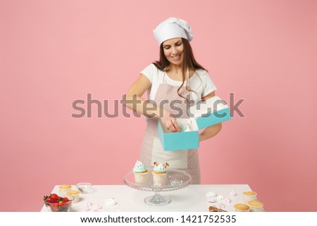 Housewife female chef cook confectioner or baker in apron white t-shirt, toque chefs hat packaging cake cupcake at table isolated on pink pastel background in studio. Mock up copy space food concept